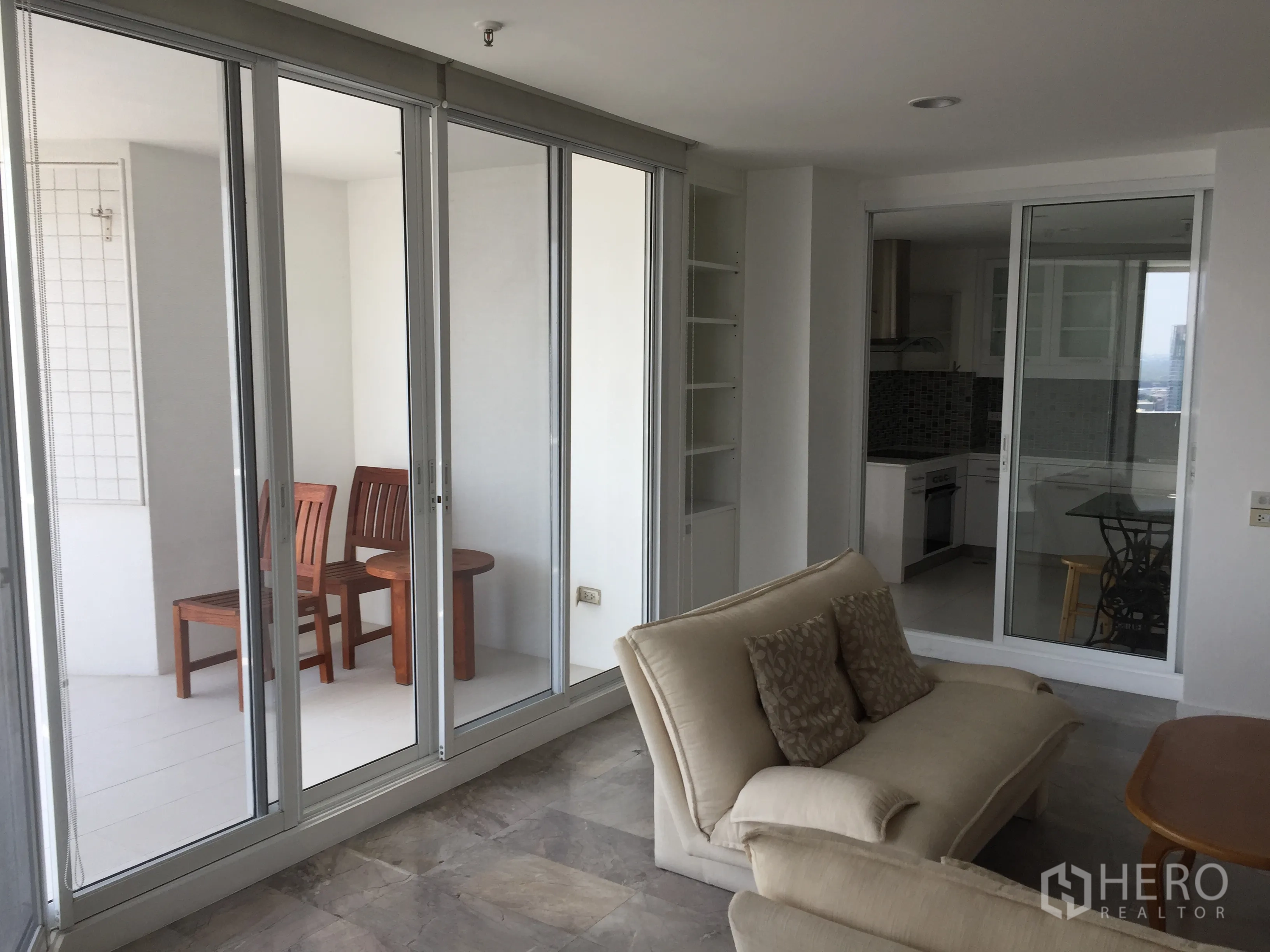 公寓 出租在Watthana, Bangkok - 客厅连接玻璃封闭阳台座位区，与独立厨房相邻。