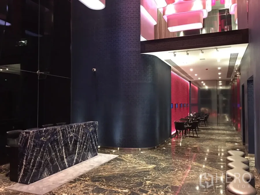 公寓 出租在Watthana, Bangkok - 公寓大堂，铺设大理石地面，深色石材前台，粉色天花灯与休息区。