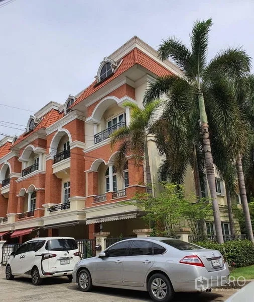 Townhouse for rent in Bueng Kum, Bangkok - Corner townhouse exterior with arched balconies, red brick facade, palm trees, and parked cars.