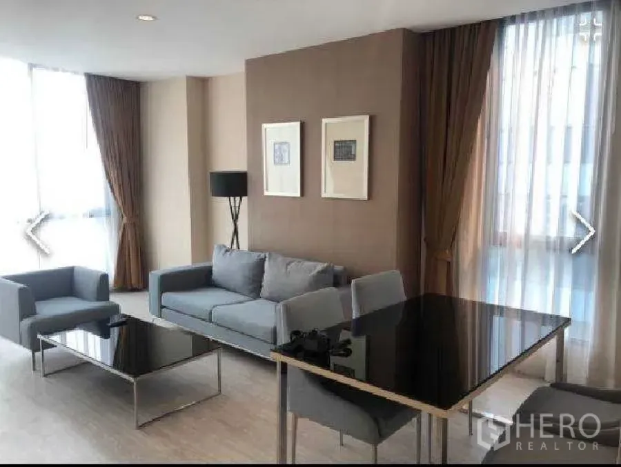 Condominium for rent in Watthana, Bangkok - Open-plan living and dining area with gray sofa, black dining table and corner windows.