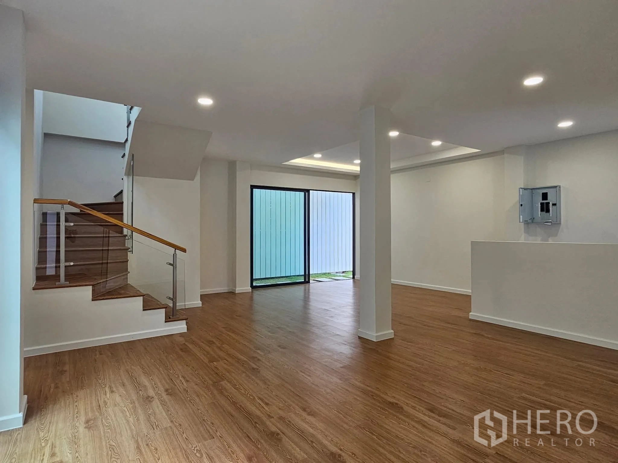 独栋房屋 for sale in Lat Phrao, Bangkok - 开放式客厅，木纹地板、筒灯照明，玻璃扶手楼梯与通向庭院的推拉门。