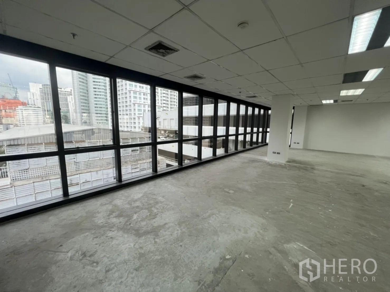 Office Space for rent in Watthana, Bangkok - Long wall of black-framed windows facing nearby towers over an unfinished concrete floor.