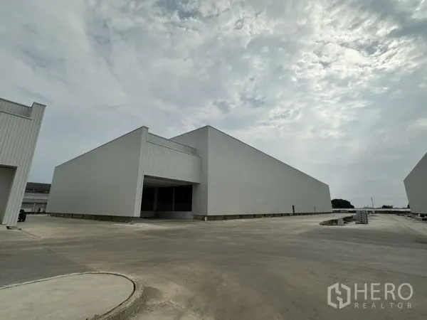 Factory for rent in Pathum Thani - New metal-clad factory warehouses with a wide concrete yard under a cloudy sky.