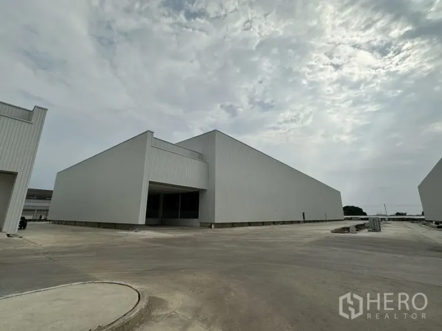 Factory for rent in Pathum Thani - New metal-clad factory warehouses with a wide concrete yard under a cloudy sky.