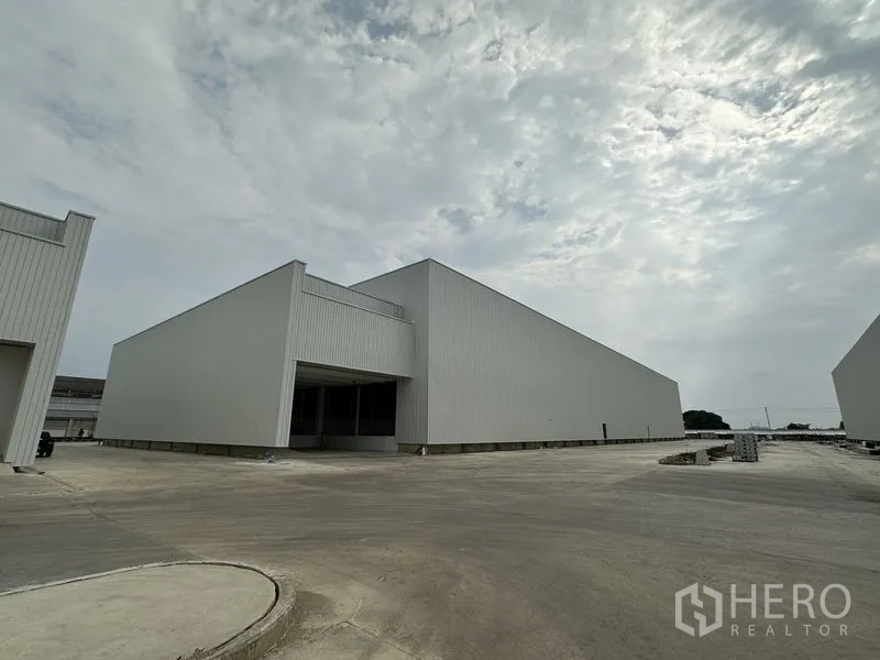 Factory for rent in Pathum Thani - New metal-clad factory warehouses with a wide concrete yard under a cloudy sky.