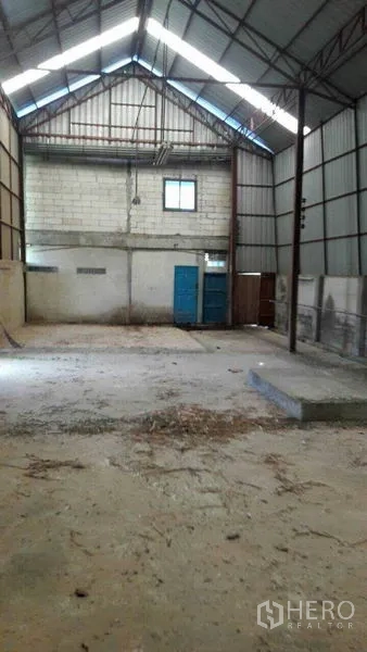 Warehouse for sale in Pathum Thani - Interior of Lam Luk Ka warehouse with high gable roof, steel trusses, concrete floor and blue metal door.