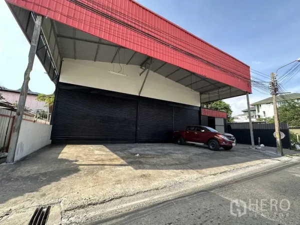 Warehouse for rent in Lat Phrao, Bangkok - Street view of Lat Phrao warehouse with red facade, wide covered loading bay and multiple black roller shutters.