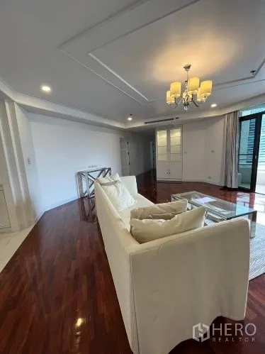 Condominium for rent in Khlong Toei, Bangkok - Bright living room with white sofa, chandelier ceiling and polished wood floors opening to a balcony in a high-rise condo.