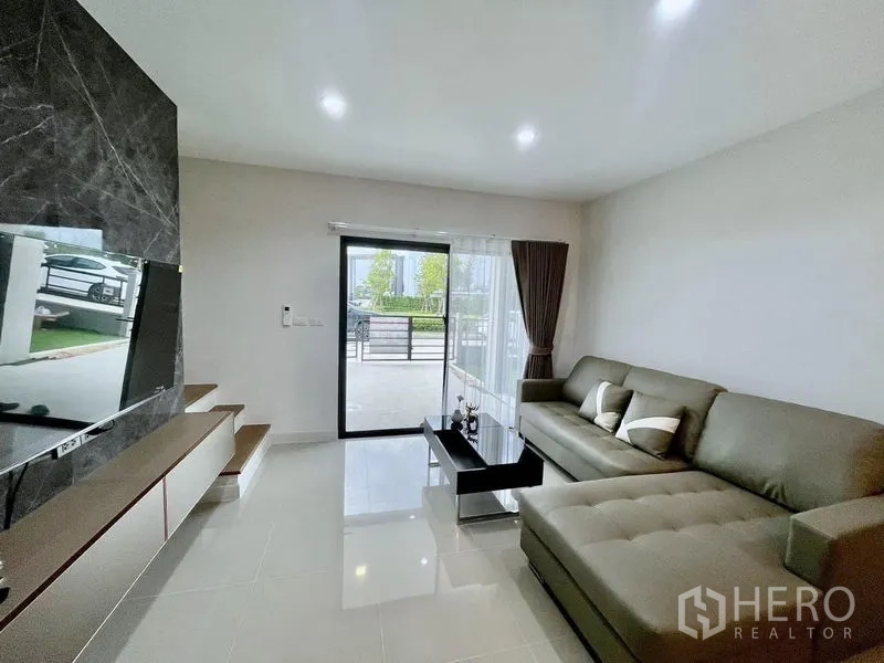 联排别墅 出租在Bang Phli, Samut Prakan - 舒适客厅，转角沙发与玻璃推拉门，连接带门禁的前露台。