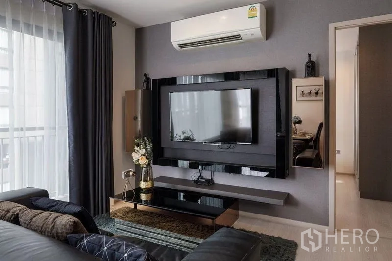 Condominium for rent in Khlong Toei, Bangkok - Living room with mounted TV, air conditioner, coffee table, and balcony window with curtains.