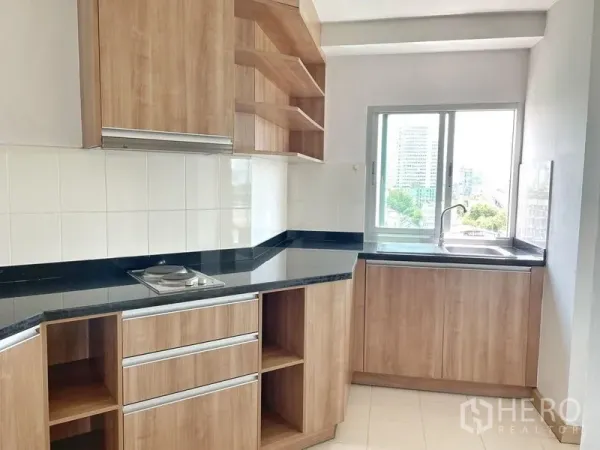 Condominium for rent in Khlong Toei, Bangkok - Bright U-shaped kitchen with wood cabinetry, black granite countertop, window and electric cooktop.