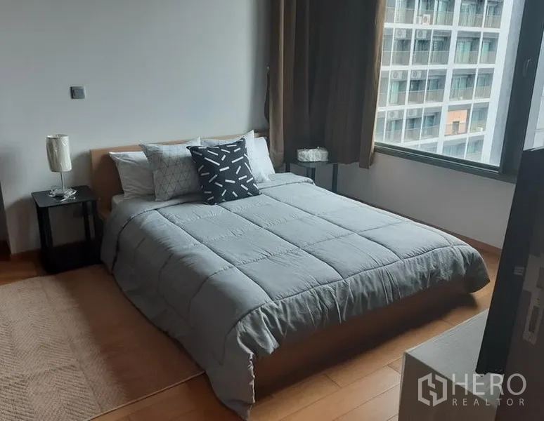 Condominium for rent in Khlong Toei, Bangkok - Cozy bedroom with double bed, bedside tables, wood flooring and a large window facing nearby buildings.