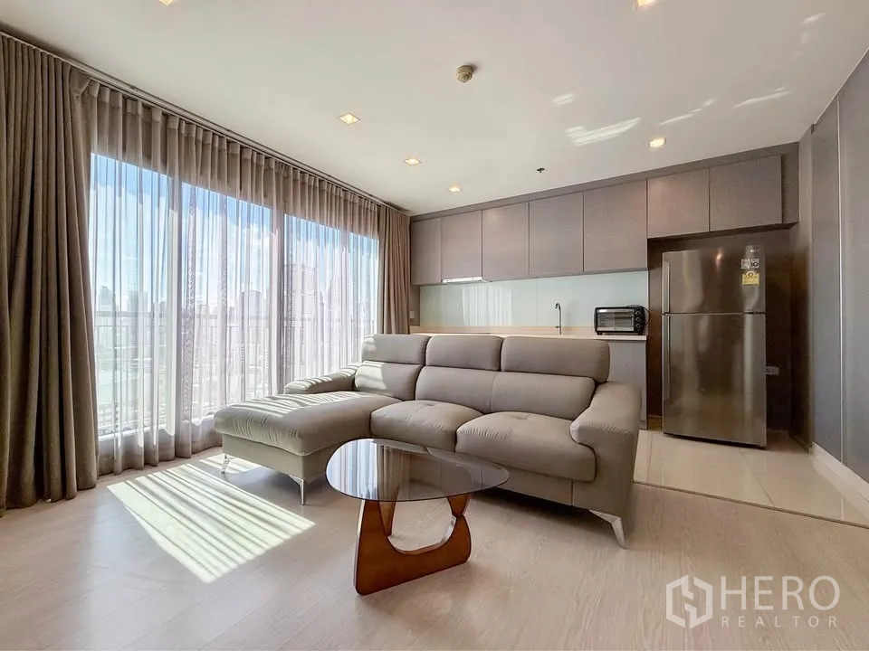 คอนโด for rent in Khlong Toei, Bangkok - พื้นที่นั่งเล่นรับแสงธรรมชาติ มีโซฟาเข้ามุม โต๊ะกลาง และครัวโทนเทา.