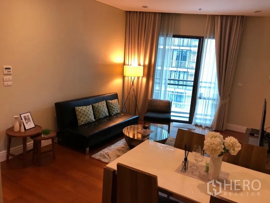 Condominium for rent or sale in Khlong Toei, Bangkok - Living room with floor-to-ceiling curtains, balcony doors, sofa and dining table under warm lighting.