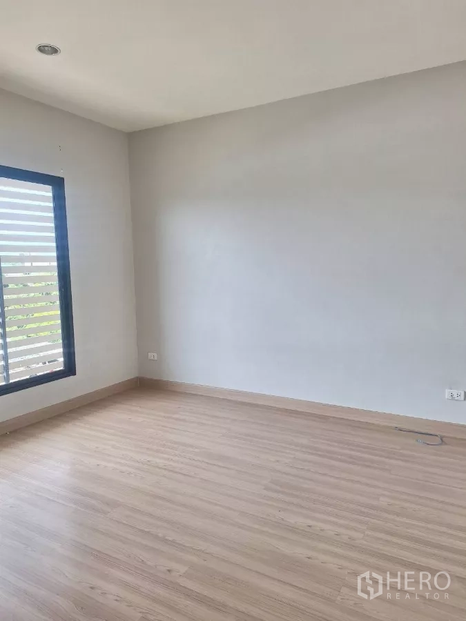 Townhouse for rent in Mueang Samut Prakan, Samut Prakan - Empty bedroom with light wood floors and a window with horizontal slats.