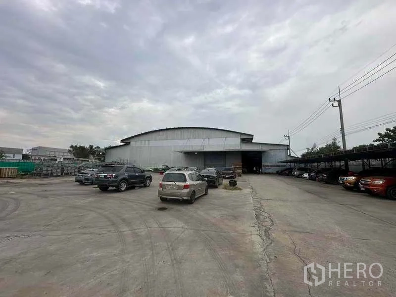 Factory for rent in Pathum Thani - Front approach to the warehouse showing loading bays, parked cars and roadside power lines.