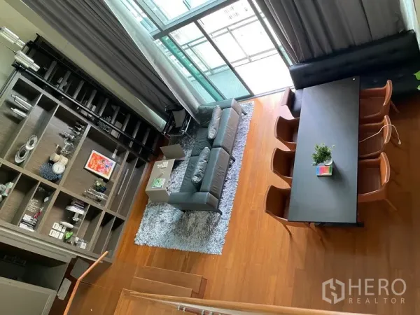 Condominium for rent in Khlong Toei, Bangkok - Aerial view of double-height living and dining area with floor-to-ceiling windows, gray sofa, and a long dining table on warm wood floors.