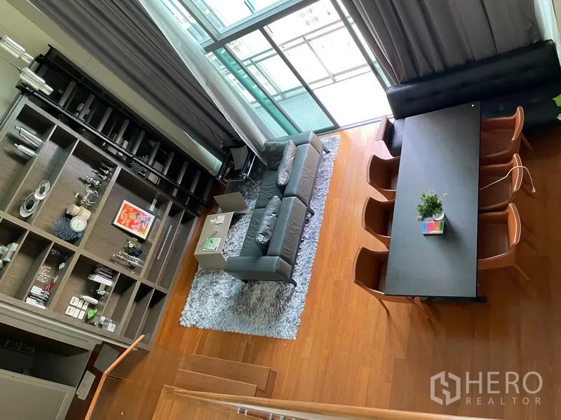 Condominium for rent in Khlong Toei, Bangkok - Aerial view of double-height living and dining area with floor-to-ceiling windows, gray sofa, and a long dining table on warm wood floors.