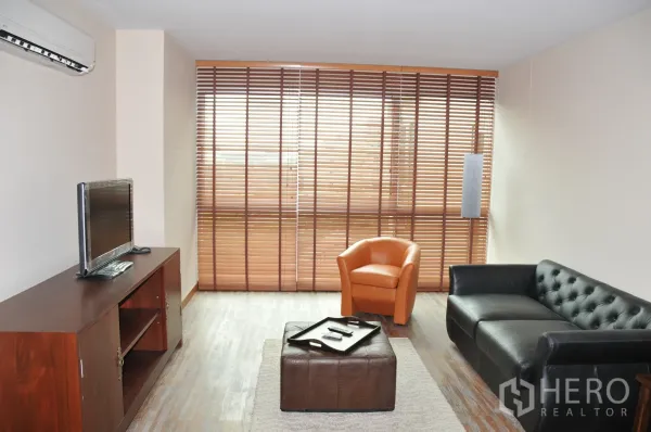 Condominium for rent in Khlong Toei, Bangkok - Living area with TV console, air conditioner, and floor-to-ceiling windows with blinds.