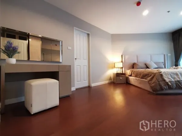 Condominium for rent or sale in Huai Khwang, Bangkok - Spacious master bedroom with upholstered bed, dressing table, ottoman, and warm lighting on wood floors.