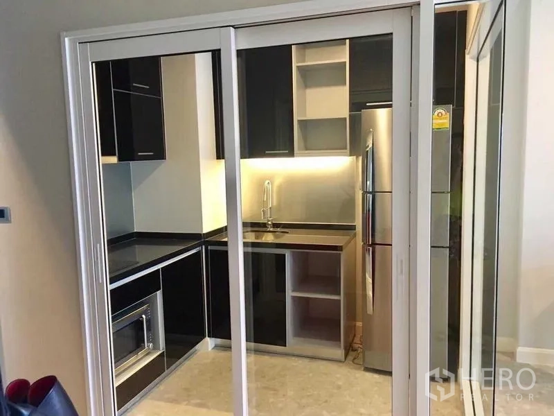 Condominium for rent in Khlong Toei, Bangkok - Compact enclosed kitchen with black cabinetry, fridge, sink and built-in microwave behind sliding glass doors.