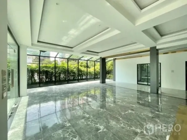 Detached House for rent or sale in Khlong Toei, Bangkok - Sunlit open-plan living area with floor-to-ceiling glass, coffered ceilings and polished marble floors facing a green hedge.