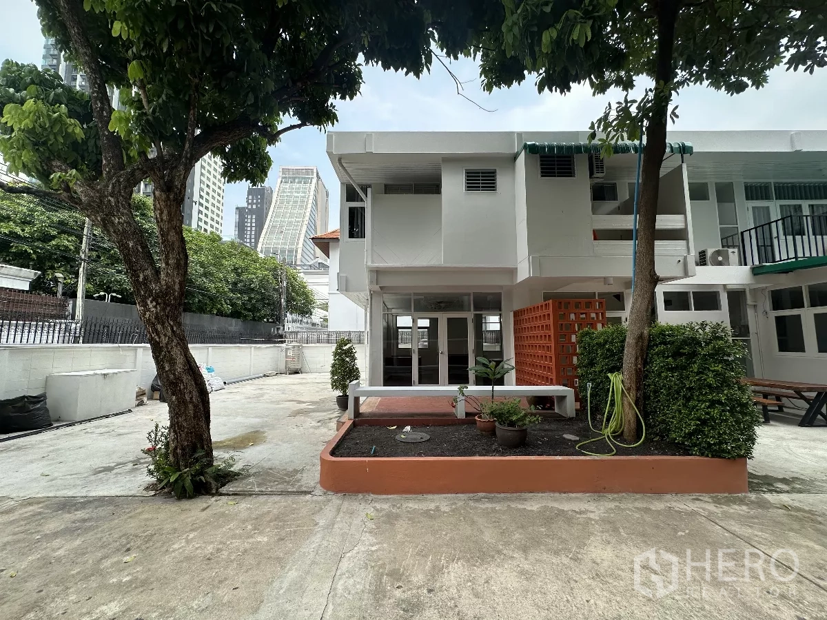 带办公室的家 出租在Watthana, Bangkok - 两层白色居家办公楼，带花坛与成树，背景为城市天际线。