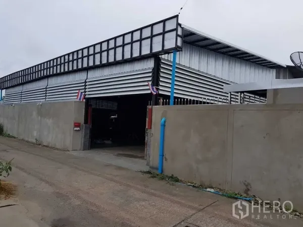 Warehouse for sale in Lam Luk Ka, Pathum Thani - Wide exterior of a metal-clad warehouse with the main gate open, blue drainage pipe and high boundary wall.