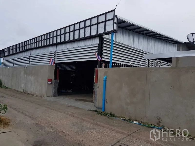 Warehouse for sale in Lam Luk Ka, Pathum Thani - Wide exterior of a metal-clad warehouse with the main gate open, blue drainage pipe and high boundary wall.