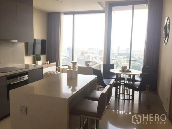 Condominium for rent in Watthana, Bangkok - Open‑plan kitchen with island, TV wall, dining set and large windows showcasing a city skyline view.