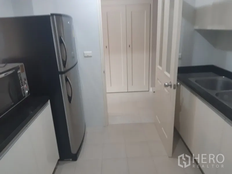 Condominium for rent in Khlong Toei, Bangkok - Kitchen view showing double-door refrigerator, microwave, and twin sinks opening to the hallway.
