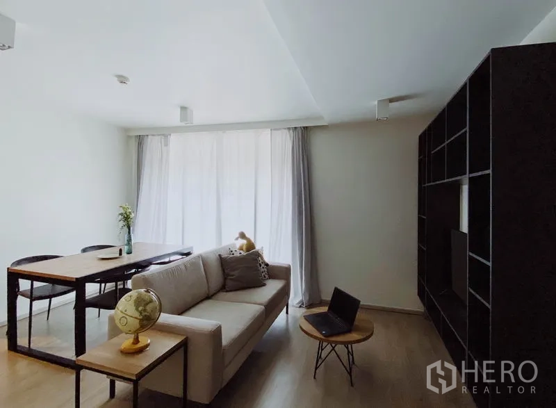 Condominium for rent in Pathum Wan, Bangkok - Minimalist living room with sofa and dining set facing a dark open-shelf media wall in soft daylight.