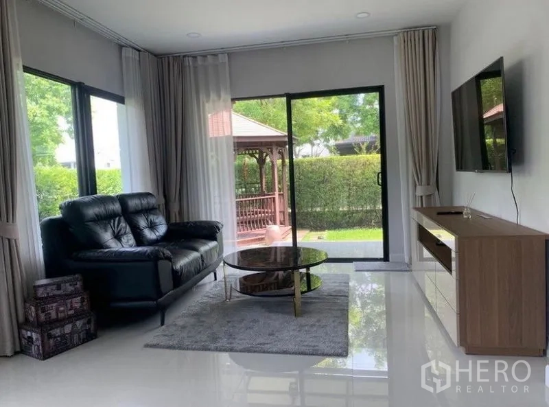 独立式住宅 出租在Prawet, Bangkok - 客厅设皮沙发与电视，推拉门通向草坪和花园凉亭。