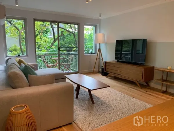 Condominium for sale in Watthana, Bangkok - Spacious living room with TV console, L-shaped sofa and large windows to a green view.