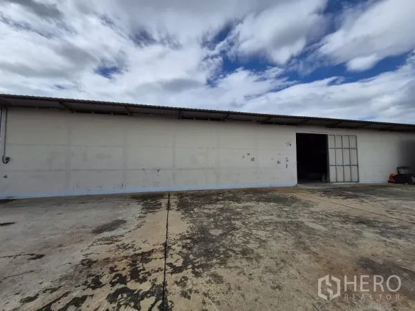 Warehouse for rent in Mueang Pathum Thani, Pathum Thani - Long single-storey warehouse exterior with sliding metal door and wide concrete loading yard under a cloudy sky.