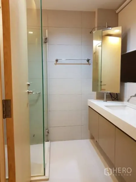 Condominium for rent in Khlong Toei, Bangkok - Modern bathroom with glass-enclosed shower, long vanity countertop, and mirror storage.