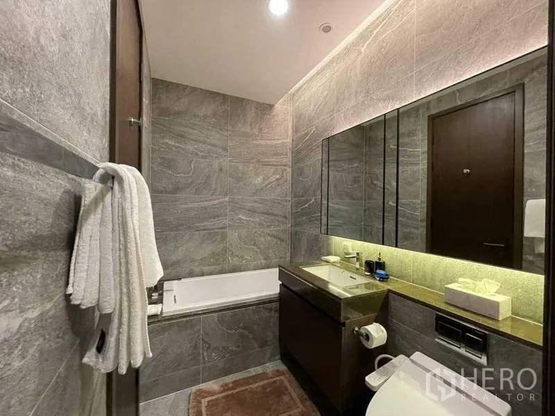 Condominium for rent in Khlong Toei, Bangkok - Bathroom with a built‑in bathtub, stone wall tiles and an illuminated vanity unit.