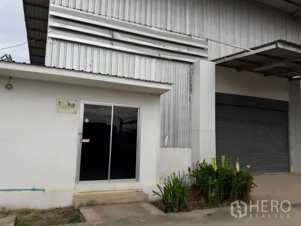 Warehouse for rent or sale in Khlong Luang, Pathum Thani - Warehouse entrance with sliding glass office door, loading roller shutter and a small garden bed.