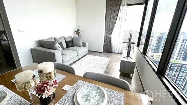 公寓 出租在Watthana, Bangkok - 四人餐桌面向灰色沙发与电视，大幅窗户呈现全景城市风光。