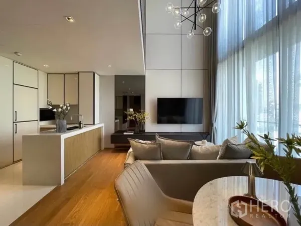 Condominium for rent in Khlong Toei, Bangkok - High-ceiling living room with sofa, TV wall and modern chandelier beside the bright kitchen.