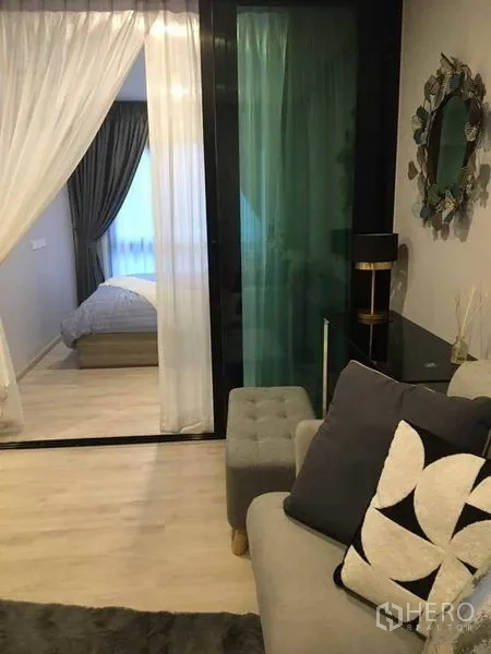 公寓 出售在Bang Na, Bangkok - 客厅配灰色沙发与脚凳，推拉玻璃隔断通向带纱帘的卧室。