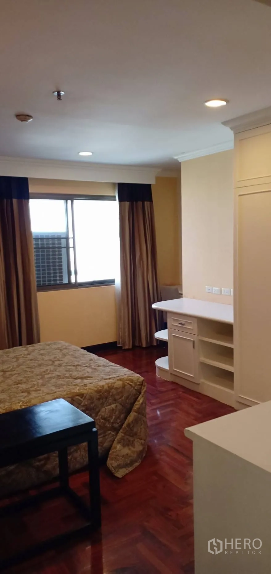 Condominium for rent in Watthana, Bangkok - Bedroom view showing built-in desk and shelves, large window, parquet flooring and soft ceiling lights.
