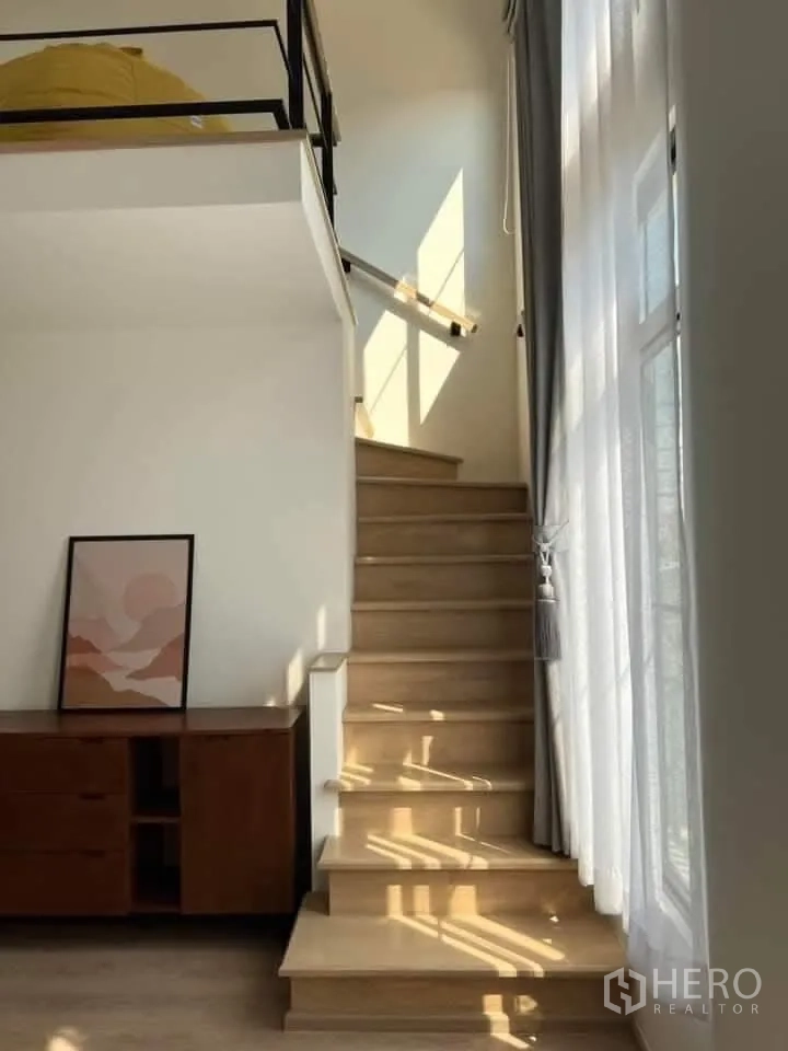 Townhouse for rent in Bang Phli, Samut Prakan - Sunlit wooden staircase beside tall curtained windows and a low console cabinet.