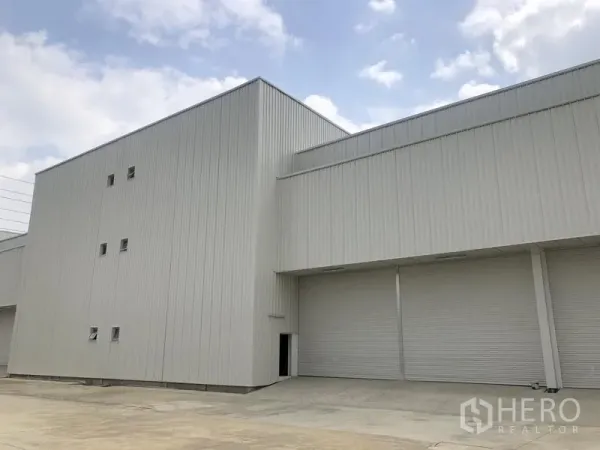 Factory for rent in Khlong Luang, Pathum Thani - Modern white steel factory exterior with multiple roller shutter doors and a wide loading forecourt under a bright sky.