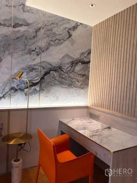 Condominium for rent in Khlong Toei, Bangkok - Compact study nook with marble-top desk, orange chair, ribbed wall paneling and side table.