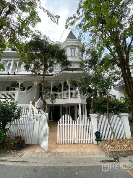 Townhouse for rent or sale in Mueang Samut Prakan, Samut Prakan - White Victorian-style townhouse facade with picket fence and mature trees.