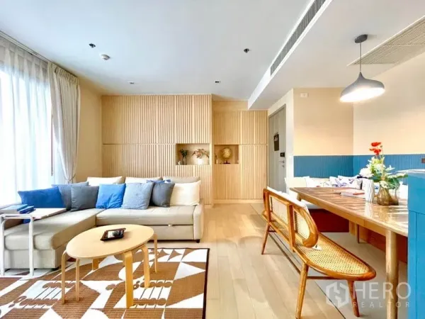 Condominium for rent or sale in Watthana, Bangkok - Airy living area with L‑shaped sofa, blue cushions, slatted wood feature wall and dining set.