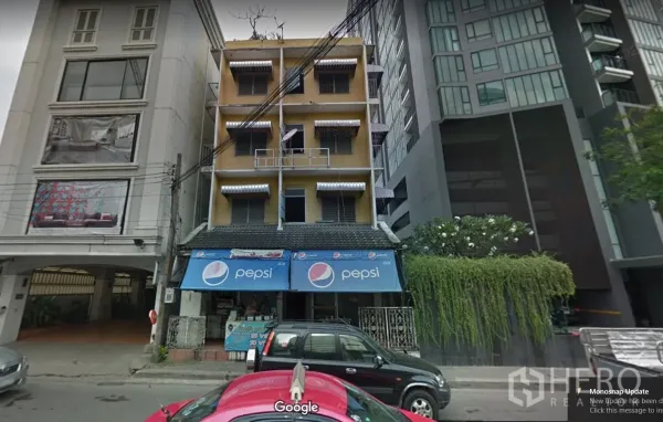 Condominium for rent in Pathum Wan, Bangkok - Street view of a low-rise shophouse with blue awnings beside a modern high-rise condominium, greenery, and city traffic near BTS Ratchadamri in Pathum Wan, Bangkok.