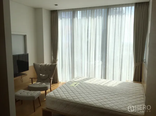Condominium for rent in Bang Rak, Bangkok - Bright bedroom with large windows, armchair and TV on a light wood floor.
