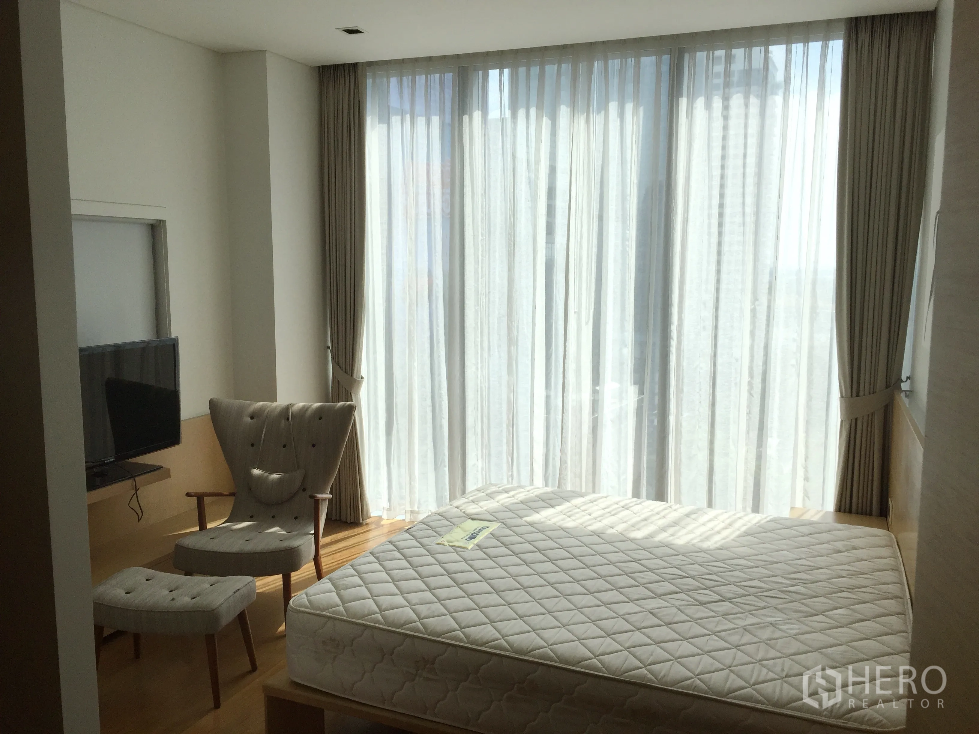 Condominium for rent in Bang Rak, Bangkok - Bright bedroom with large windows, armchair and TV on a light wood floor.
